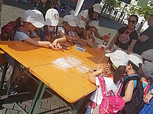 Kinder mit weißer Kappe an einem großen Tisch auf dem verschiedenfarbiges Wasser in Schalen und Pipetten liegen