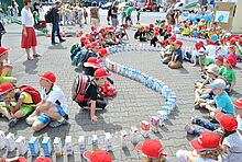 Viele Kinder mit roten Kappen bauen eine Domino-Schlange aus Tetra-Paks.